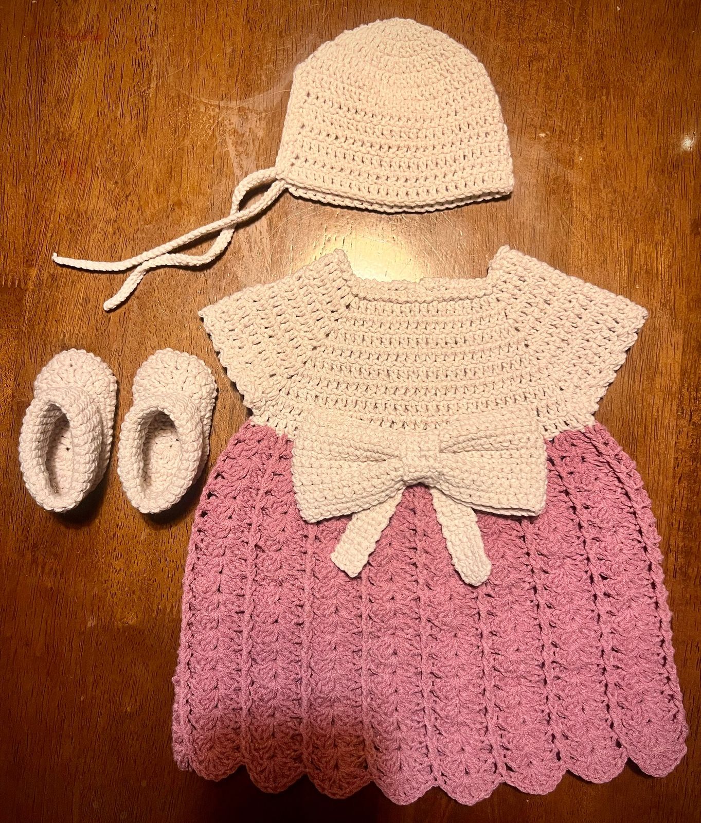 A crocheted baby gift set including a bonnet, dress and booties in cream and dusty pink, laid flat on a wooden table.