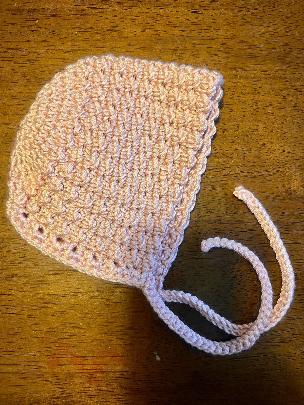 A hand-crocheted pink baby bonnet laid flat on a wooden table.