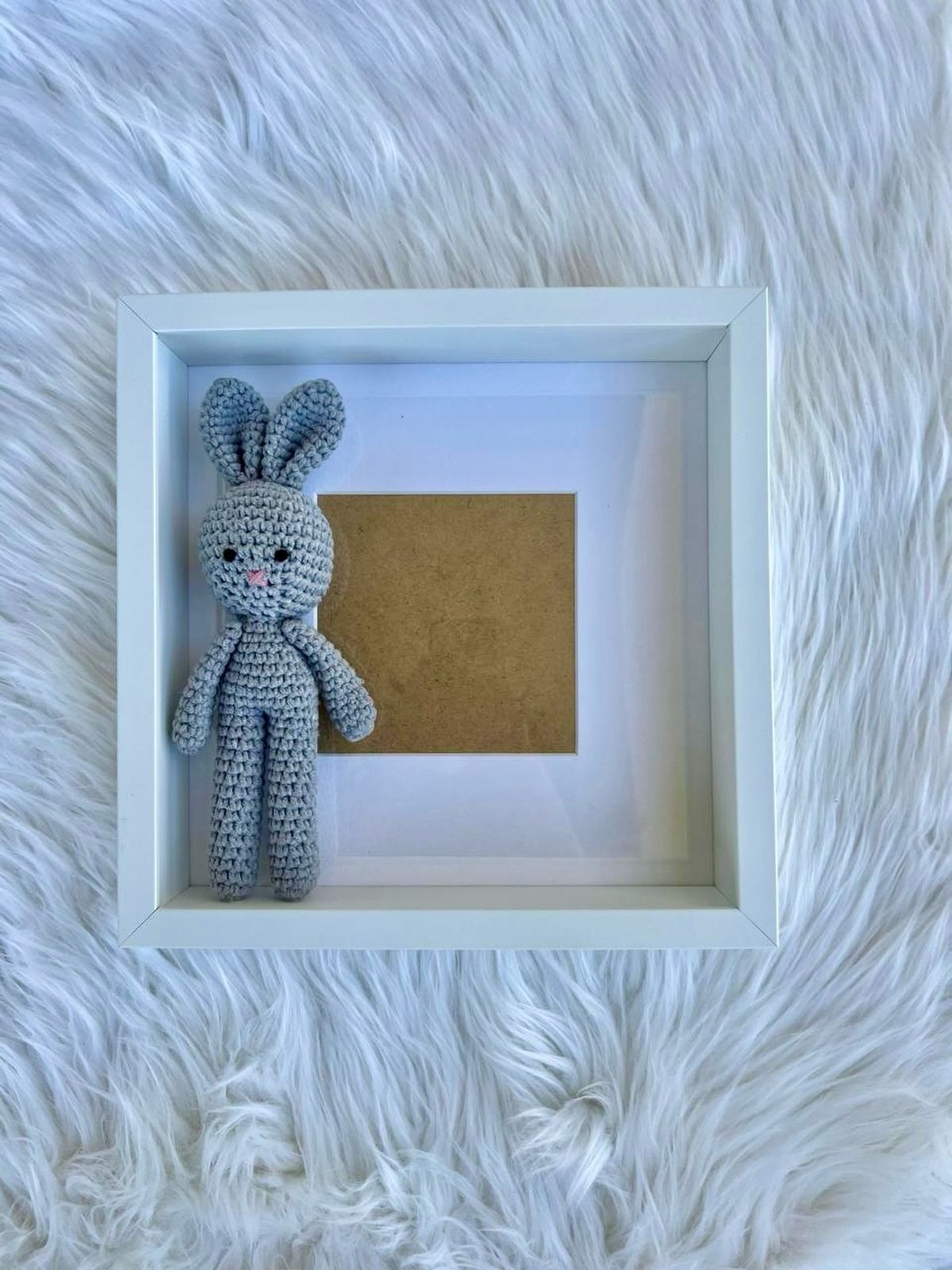 A small grey crochet bunny propped in a white square photo frame on a fur background.