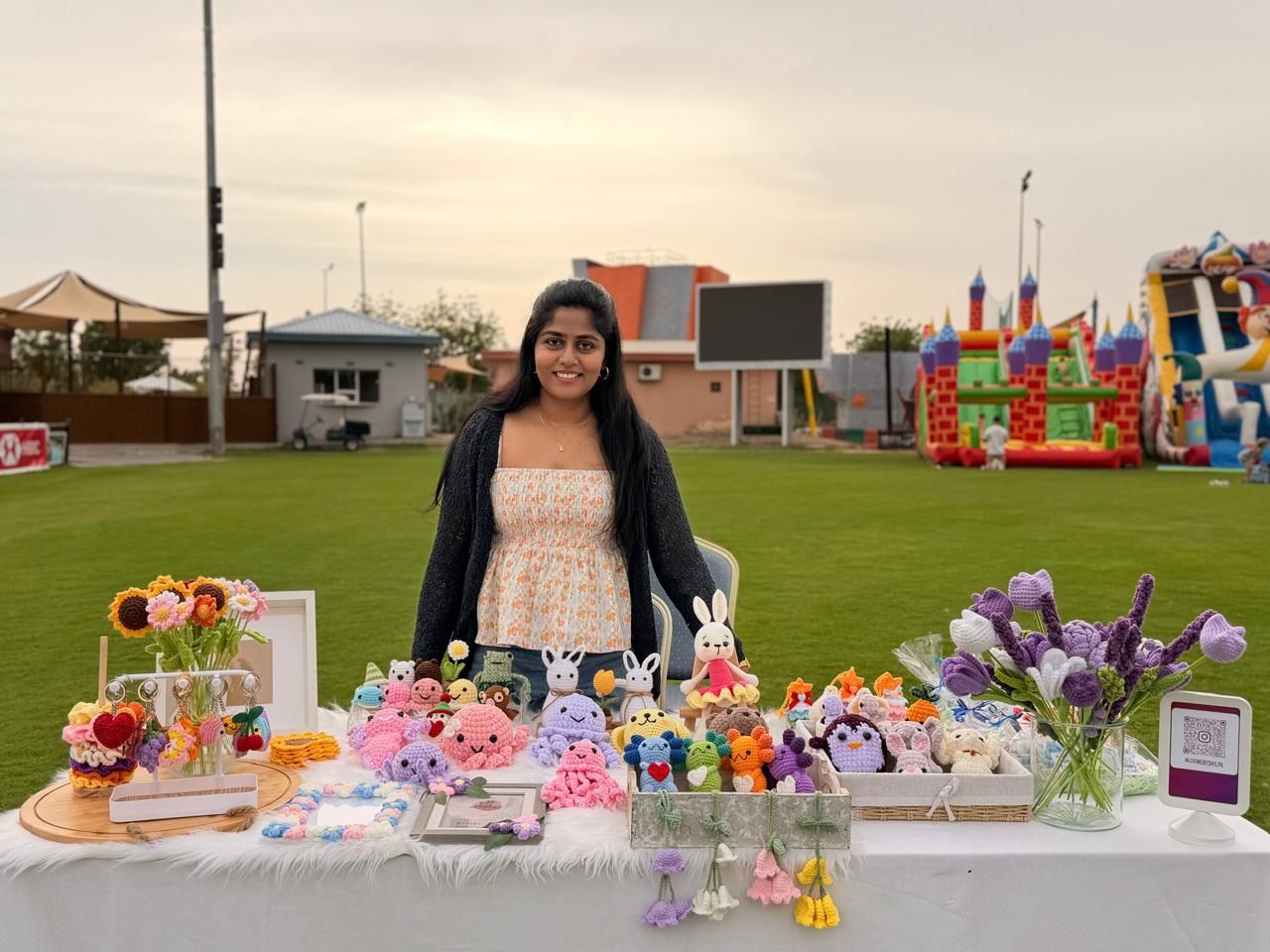 Shilpa standing behind her market stall table, neatly arranged with crochet flowers, bouquets and amigurumi, on a grass field in Al Ain.
